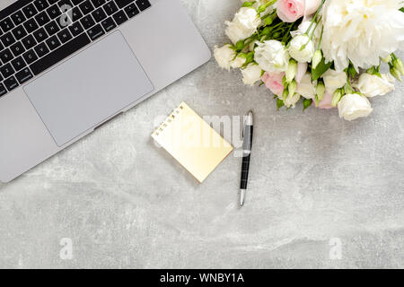 Frau Büro Schreibtisch Tisch mit Laptop Computer, Blumen, gelber Notizblock, Schreibwaren auf Beton Stein Hintergrund. Flach, Ansicht von oben feminine Arbeitsbereich Stockfoto