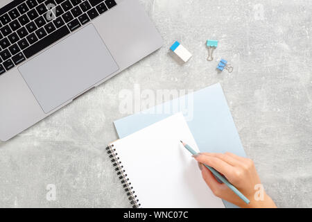 Flach, Ansicht von oben Bürotisch Schreibtisch. Mit Laptop, Hand das Schreiben von Text in Tagebuch auf Beton Stein Arbeitsbereich. Feminine Arbeitsbereich Konz Stockfoto