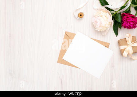 Flach Päonien Blüten, Handwerk Umschlag, leeres Papier Karte, Geschenkbox, Band auf Holz- Hintergrund. Ansicht von oben feminine Arbeitsbereich, Einladung Hochzeit Auto Stockfoto