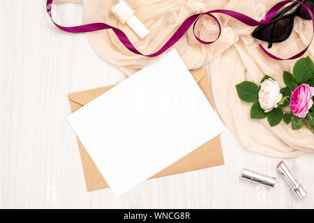 Flach leere weiße Grußkarte mit craft-Umschlag, Rosen Blumen, Lippenstift, Band auf Holz- Hintergrund. Ansicht von oben feminine home office Schreibtisch Stockfoto