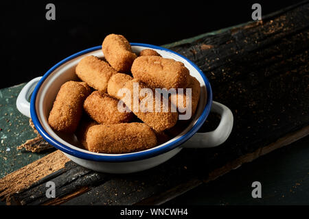 Nahaufnahme von einigen spanischen Kroketten in weiß und blau Emaille Teller auf einem rustikalen Dunkelgrau Holztisch, auf schwarzem Hintergrund Stockfoto