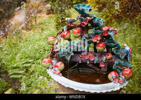 Dekorative Dekoration des Gartens. Keramik Topf mit einer Statue von einem Apfelbaum mit roten Äpfeln und einem kleinen bunten Vogel. grünes Gras im Bac Stockfoto