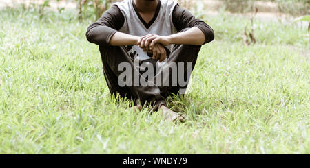 Close up Beschnittenes Bild des jungen Mannes in Freizeitkleidung sitzt über grüne Gras in dem Park am Morgen Zeit des Tages. Vorderansicht. Das Konzept der Ausgaben le Stockfoto
