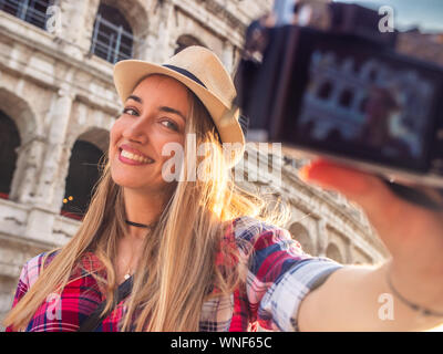 Junge blonde Frau mit blauen Augen und langen Haaren, wobei selfie mit einem Vintage Kamera am Kolosseum, Rom. Großen Hut, rot kariert Hemd. Stockfoto