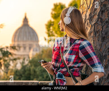 Schöne, junge blonde Frau mit langen Haaren das Hören von Musik mit weißen headpones und Smartphone. Lehnte sich an einen Baum. Rom, Italien, Sonnenuntergang. Saint Stockfoto