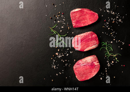 Rohes Rindfleisch Steaks mit Salz, Pfeffer und Rosmarin, geschossen von oben auf einem schwarzen Hintergrund mit Copyspace Stockfoto