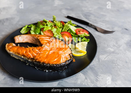Gebratener Lachs Fisch Steak mit Gemüse und Salat Zitrone auf schwarze Platte. Kreative Gestaltung aus Fisch, Salat, Tomate, Gurke. Gesunde Ernährung Konzept Stockfoto