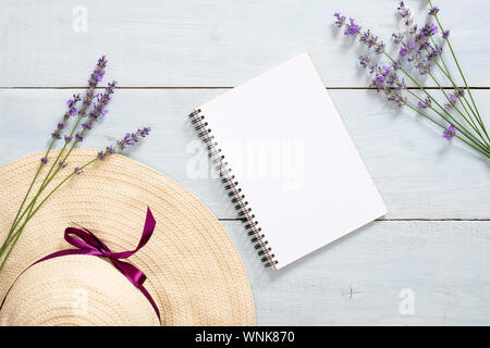 Minimale home office Schreibtisch mit leeren Notebook, Strohhut und Lavendelblüten auf rustikalen blau Holz- Hintergrund. Flach, Ansicht von oben Mock up. Stockfoto