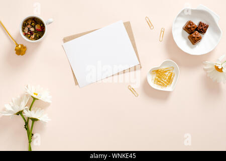 Flach Komposition mit leere weiße Karte, Handwerk Papier Umschlag, Kamillenblüten, goldene Schreibwaren, Tasse Tee und Süßigkeiten auf Pastell rosa Hintergrund. T Stockfoto