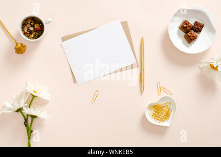 Flach Komposition mit leere weiße Karte, Handwerk Papier Umschlag, Kamillenblüten, goldene Schreibwaren, Tasse Tee und Süßigkeiten auf Pastell rosa Hintergrund. T Stockfoto