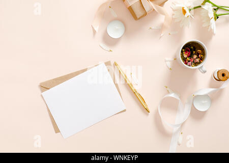 Romantische flatlay Komposition mit leerem Papier Karte Mockup, Umschlag, Kräutertee, Band, Daisy Kamille Blüte Pastell rosa Hintergrund. Feminine ho Stockfoto