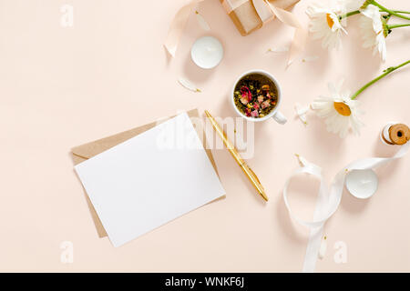 Romantische flatlay Komposition mit leerem Papier Karte Mockup, Umschlag, Kräutertee, Band, Daisy Kamille Blüte Pastell rosa Hintergrund. Feminine ho Stockfoto