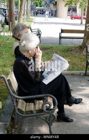 Elegante schöne ältere Frau tragen altmodische Kleid, sitzen auf einer Bank in einem öffentlichen Garten, Zeitung lesen. 70, 80, 90 Jahre alte Dame Stockfoto