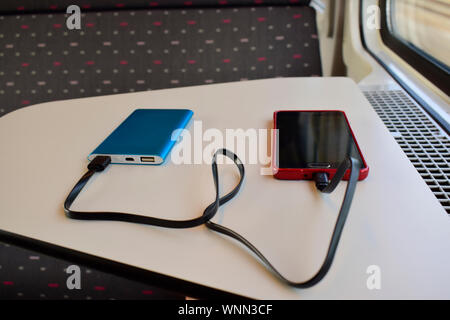 Schwarz und Rot Smartphone laden mit Blue Power Bank auf weißem Hintergrund in der Nähe des Sitzes im Zug gegen die Sonne durch das Fenster. Stockfoto