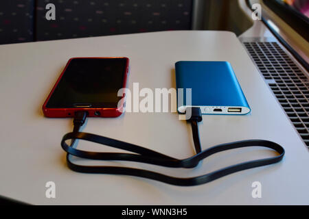 Schwarz und Rot Smartphone laden mit Blue Power Bank auf weißem Hintergrund in der Nähe des Sitzes im Zug gegen die Sonne durch das Fenster. Stockfoto