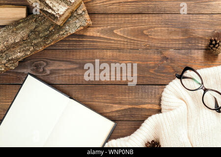 Feminine Schreibtisch mit weißen Strickschal, Gläser, Brennholz, Buch mit leeren Seiten auf Holz- Hintergrund. Hygge, Nordic, skandinavischen Stil Arbeitsplatz. Stockfoto