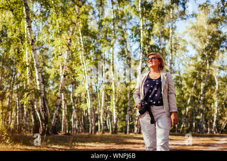 Frau mittleren Alters Aufnehmen von Bildern mit der Kamera im Herbst Wald. Stilvolle senior Frau Wandern und Genießen Hobby Fotografieren Stockfoto