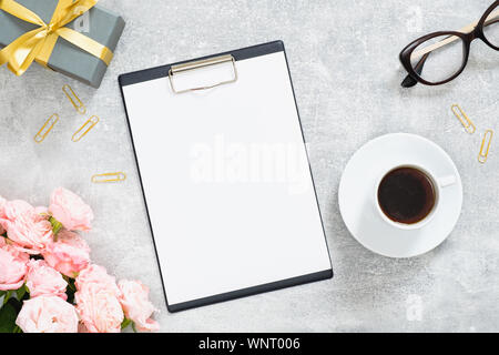 Elegante feminine Schreibtisch mit leerem Papier Zwischenablage Mockup, rosa Blumen, Geschenkbox mit gold Farbband, Tasse, Gläser auf Stein Hintergrund. Stockfoto