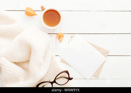 Herbst Komposition. Feminine Arbeitsbereich mit leeren Brief, Kaffee Becher, Gläser, aus Gewirken plaid auf hölzernen weißen Hintergrund. Ansicht von oben, flach, kopieren. Stockfoto