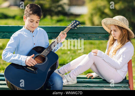 Junge Mädchen und Jungen mit der Gitarre auf der Bank sitzt im Park. Kinder lieben. Stockfoto
