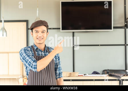 Die jungen asiatischen Mann, ist das Tragen einer Schürze selbstbewusst Lächeln, und zeigen Sie auf etwas über ihn in die Leere Monitor hinter ihm an einem Arbeitsplatz in der m Stockfoto