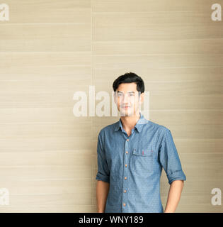 Jungen asiatischen gutaussehenden Mann, der trägt ein blaues T-Shirt Casual Wear steht vor der hölzernen Wand im Büro morgen Zeit. lifestyle Portrait. Stockfoto