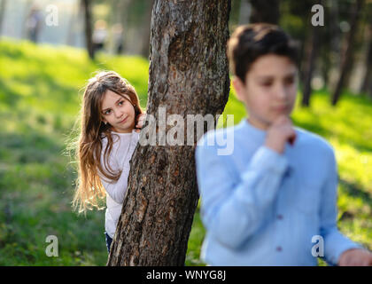 Junge und Mädchen spielen Verstecken und im Park suchen. Mädchen beobachten auf Freund. Mädchen versteckt sich hinter dem Baum und lauern auf Jungen. Stockfoto