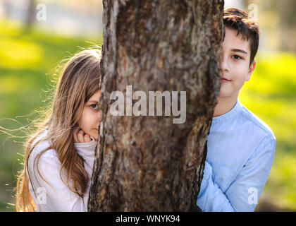 Junge und Mädchen spielen Verstecken und im Park suchen. Mädchen beobachten auf Freund. Mädchen versteckt sich hinter dem Baum und lauern auf Jungen. Stockfoto