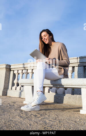 Lachende Frau mit der Tablette, während auf einer Bank sitzt Stockfoto