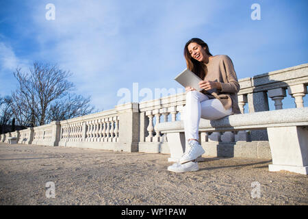 Lachende Frau mit der Tablette, während auf einer Bank sitzt Stockfoto