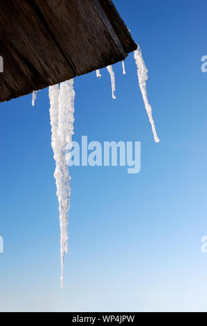 Die Eiszapfen, die von einem hölzernen Dach hängen, gegen den blauen Himmel und die Berge Stockfoto