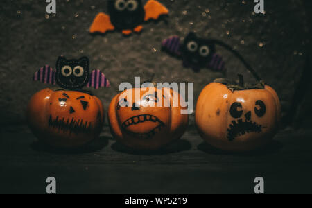 Kleine Kürbisse mit unterschiedlichen Gesichtern stehen auf einem Holzregal, im Hintergrund sind handgefertigt Fledermäuse aus Filz. Halloween Attribute Stockfoto