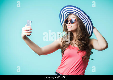 Cute brunette Mädchen nimmt einen selfie. Sie lächelt auf blauem Hintergrund. Gerne Frau trägt Hut und Rosa oben Gestreifte Stockfoto