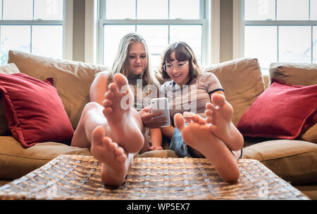 Zwei Mädchen sitzen auf der Couch mit den Füßen bis auf Handy. Stockfoto
