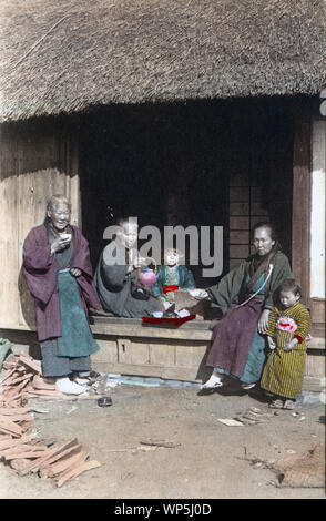 [1900s Japan - Japanische Tee Pause im Land] - Drei ältere Frauen und zwei Kinder, die alle im Kimono, haben Kaffee auf der Veranda eines ländlichen Haus mit Strohdach. Gehackte Holz liegt rund herum. 20. jahrhundert alte Ansichtskarte. Stockfoto