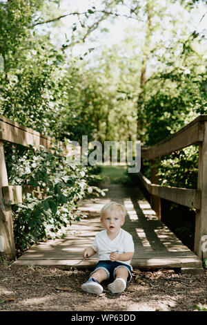 Süße kleine blonde Kind Junge Kind sitzen und Lachen vor der hölzernen Brücke über einen Bach im Park im Wald im Sommer Stockfoto