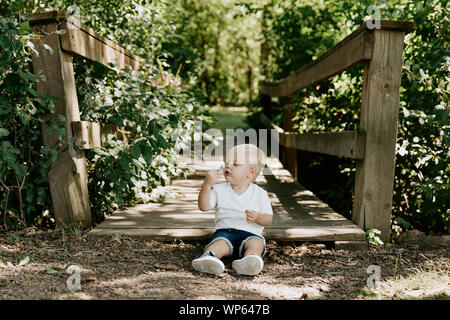 Süße kleine blonde Kind Junge Kind sitzen und Lachen vor der hölzernen Brücke über einen Bach im Park im Wald im Sommer Stockfoto