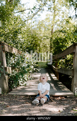 Süße kleine blonde Kind Junge Kind sitzen und Lachen vor der hölzernen Brücke über einen Bach im Park im Wald im Sommer Stockfoto