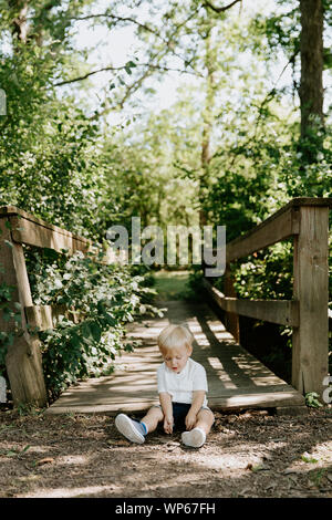 Süße kleine blonde Kind Junge Kind sitzen und Lachen vor der hölzernen Brücke über einen Bach im Park im Wald im Sommer Stockfoto