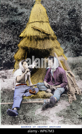 [1920s Japan - Japanische Bauern das Rauchen einer Pfeife] - ein japanischer Landwirt und sein Freund sind, das Rauchen eine kiseru Rohr während einer Pause. Sie tragen westliche Kleidung mit tabi Schuhe. Diese Postkarte ist aus einer Serie über die japanische Landwirtschaft, Landwirt das Leben in Japan. 20. jahrhundert alte Ansichtskarte. Stockfoto