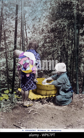 [1920s Japan - Japanische Bauer Frau Waschen] - eine Frau im Yukata und traditionellen headwear ist die Wäsche mit einem Oke (hölzerne Schaufel). Ein Mädchen mit einem Baby steht neben ihr. Diese Postkarte ist aus einer Serie über die japanische Landwirtschaft, Landwirt das Leben in Japan. 20. jahrhundert alte Ansichtskarte. Stockfoto