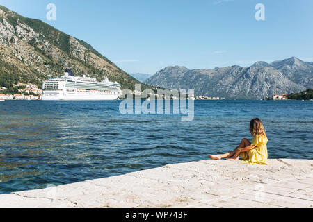 Ein Mädchen in einem gelben Kleid sitzt am Strand und blickt auf einem Segelschiff. Sie träumt, oder denkt, oder genießt die Einsamkeit. Stockfoto