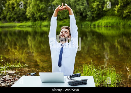 White Collar erstreckt sich beim Sitzen am Tisch. Ein Büroangestellter sitzt an einem Tisch inmitten einer natürlichen Landschaft mit einem wilden Fluss. Auf seinem Schreibtisch ist ein Erl Stockfoto