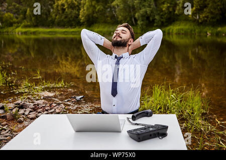 Ein Büroangestellter sips Muskeln, während er sich in einem Büro an einem Tisch mit einem Laptop und einem Festnetz Druckknopf Telefon sitzen. Büro Schreibtisch am Ufer ich Stockfoto