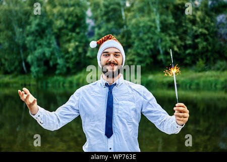Eine junge Lächeln, Geschäftsmann, tragen ein Weihnachtsmann hat, hält eine brennende Wunderkerze in der Hand und stand vor dem hintergrund der grünen Bäumen und einem Fluss Stockfoto