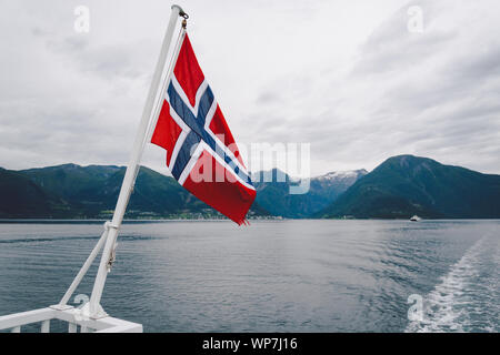 Norwegische Flagge hängen an der Reling des Schiffes und winken über dem Wasser. Norwegische Fjord mit einer Flagge. Mit der Fähre in Norwegen. Norwegen Flagge auf Meer Stockfoto