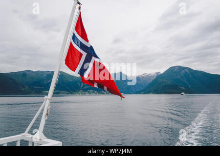 Norwegische Flagge hängen an der Reling des Schiffes und winken über dem Wasser. Norwegische Fjord mit einer Flagge. Mit der Fähre in Norwegen. Norwegen Flagge auf Meer Stockfoto