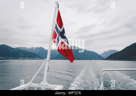 Norwegische Flagge hängen an der Reling des Schiffes und winken über dem Wasser. Norwegische Fjord mit einer Flagge. Mit der Fähre in Norwegen. Norwegen Flagge auf Meer Stockfoto