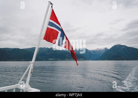 Norwegische Flagge hängen an der Reling des Schiffes und winken über dem Wasser. Norwegische Fjord mit einer Flagge. Mit der Fähre in Norwegen. Norwegen Flagge auf Meer Stockfoto