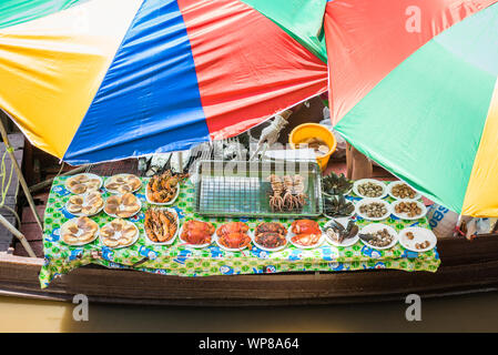 Amphawa, Samut Songkhram/Thailand - Juni 15, 2019: Tabelle mit gegrillten Meeresfrüchten unter bunten Sonnenschirmen in einem Boot an schwimmenden Markt Amphawa. Stockfoto